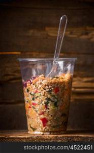 Healthy bulgur salad in plastic cup with plastic fork on dark wooden background. Take away lunch