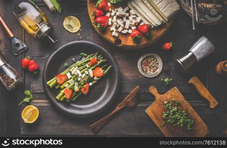 Healthy dietary lunch dish with green asparagus and diced feta cheese, herbs and strawberries on black plate. Rustic wooden background. Concept for a tasty and healthy vegetarian home cooking meal.