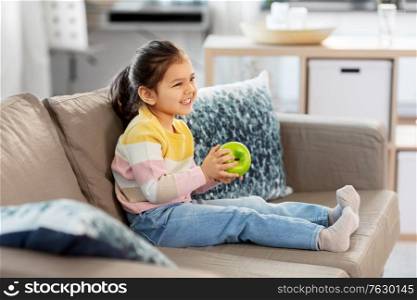 healthy eating and childhood concept - happy smiling little girl with green apple sitting on sofa at home. happy little girl with apple sitting on sofa