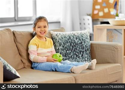 healthy eating and childhood concept - happy smiling little girl with green apple sitting on sofa at home. happy little girl with apple sitting on sofa