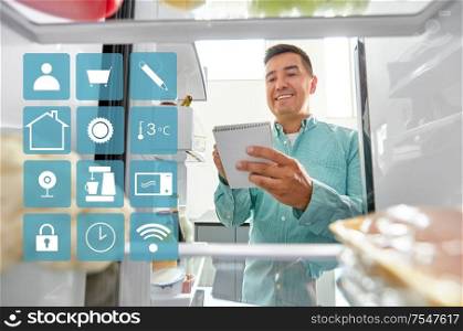 healthy eating and diet concept - smiling man opening fridge and making list of necessery food at kitchen and white smart home app icons over blue. man making list of necessary food at home fridge