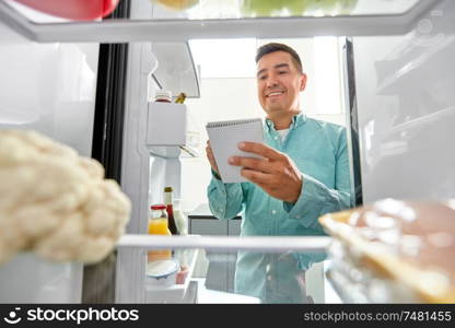 healthy eating and diet concept - smiling middle-aged man opening fridge and making list of necessary food at home kitchen. man making list of necessary food at home fridge