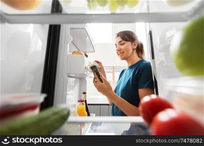 healthy eating and diet concept - woman at fridge with smartphone making list of necessary food at home kitchen. woman with smartphone makes list of food in fridge