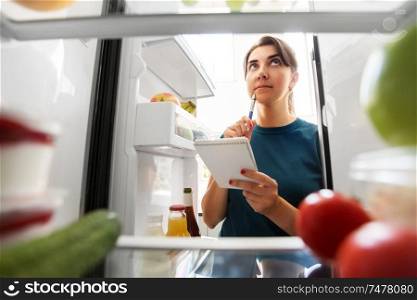 healthy eating and diet concept - woman opening fridge and making list of necessary food at home kitchen. woman making list of necessary food at home fridge