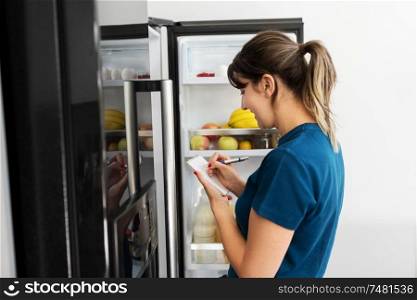 healthy eating and diet concept - woman opening fridge and making list of necessary food at home kitchen. woman making list of necessary food at home fridge