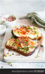 Healthy eating concept. Toast with avocado cream and smoked salmon on the white wooden board. Smoked salmon, cream cheese and pesto toast sandwiches with radish microgreens sprouts.