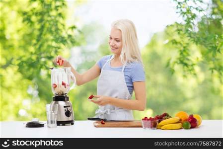 healthy eating, cooking, food and people concept - happy smiling young woman putting fruits and berries for fruit smoothie to blender shaker over green natural background. smiling woman with blender making fruit smoothie