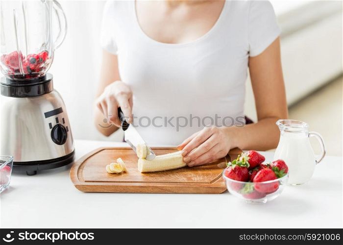 healthy eating, cooking, vegetarian food, dieting and people concept - close up of young woman with blender chopping banana for fruit shake at home