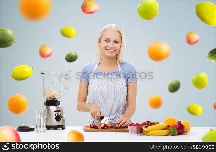 healthy eating, cooking, vegetarian food, dieting and people concept - smiling young woman with blender chopping fruits and berries for fruit shake over gray background with falling vegetables