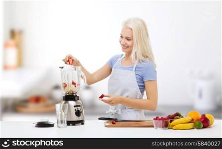 healthy eating, cooking, vegetarian food, dieting and people concept - smiling young woman putting fruits and berries for fruit shake to blender shaker over kitchen background