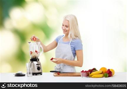 healthy eating, cooking, vegetarian food, dieting and people concept - smiling young woman putting fruits and berries for fruit shake to blender shaker over green natural background