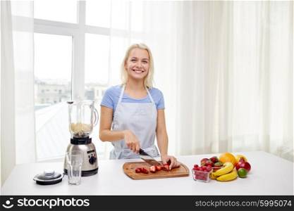 healthy eating, cooking, vegetarian food, dieting and people concept - smiling young woman with blender chopping fruits and berries for fruit shake at home
