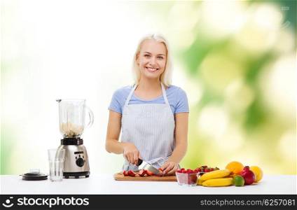 healthy eating, cooking, vegetarian food, dieting and people concept - smiling young woman with blender chopping fruits and berries for fruit shake over green natural background