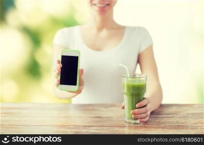 healthy eating, diet, detox, technology and people concept - close up of woman with smartphone green juice sitting at wooden table over green natural background