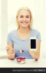 healthy eating, dieting and people concept - smiling young woman eating fruit salad and showing blank smartphone screen at home