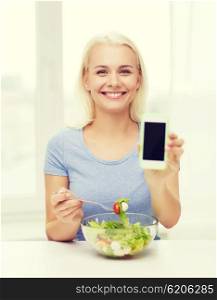 healthy eating, dieting and people concept - smiling young woman eating vegetable salad and showing blank smartphone screen at home