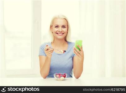 healthy eating, dieting and people concept - smiling young woman with smartphone eating fruit salad at home