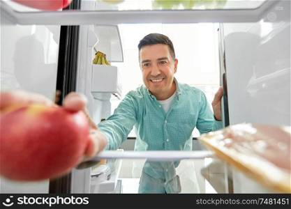 healthy eating, food and diet concept - happy middle-aged man taking apple from fridge at home kitchen. man taking apple from fridge at home kitchen