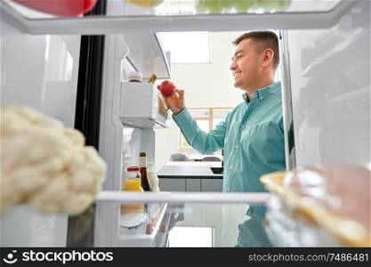 healthy eating, food and diet concept - happy middle-aged man taking apple from fridge at home kitchen. man taking apple from fridge at home kitchen