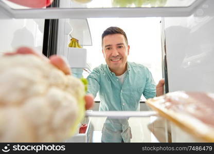 healthy eating, food and vegetable diet concept - happy middle-aged man taking cauliflower from fridge at home kitchen. man taking vegetable from fridge at home kitchen