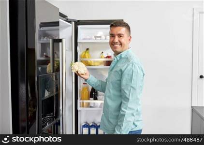 healthy eating, food and vegetable diet concept - happy middle-aged man taking cauliflower from fridge at home kitchen. man taking vegetable from fridge at home kitchen