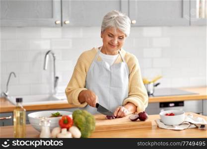 healthy eating, food cooking and culinary concept - happy smiling senior woman with knife chopping red onion on kitchen at home. happy woman chopping red onion on kitchen