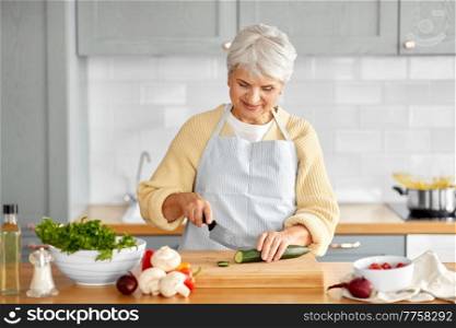 healthy eating, food cooking and culinary concept - happy smiling senior woman with knife chopping cucumber on kitchen at home. happy woman chopping cucumber on kitchen