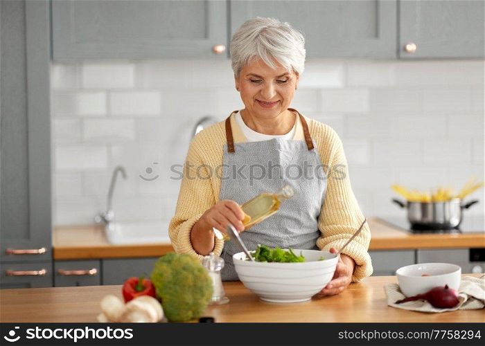 healthy eating, food cooking and culinary concept - happy smiling senior woman with olive oil making vegetable salad on kitchen at home. happy woman adding cooking oil to salad on kitchen