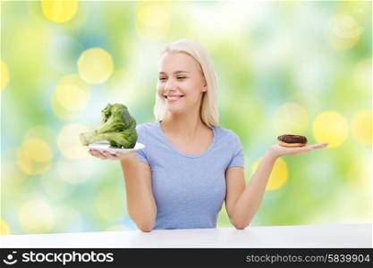healthy eating, junk food, diet and choice people concept - smiling woman choosing between broccoli and donut over summer green holidays lights background
