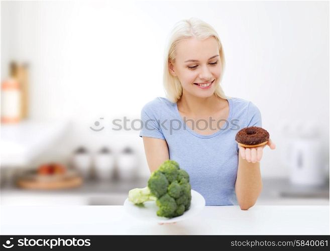 healthy eating, junk food, diet and choice people concept - smiling woman choosing between broccoli and donut over kitchen background