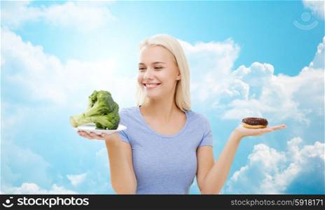 healthy eating, junk food, diet and choice people concept - smiling woman choosing between broccoli and donut over blue sky and clouds background