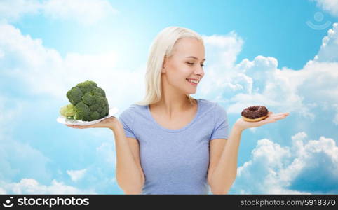 healthy eating, junk food, diet and choice people concept - smiling woman choosing between broccoli and donut over blue sky and clouds background