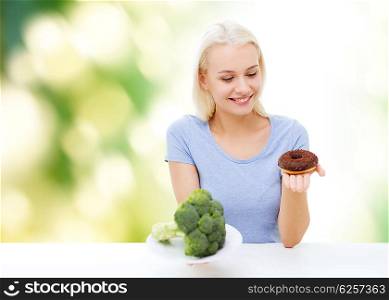 healthy eating, junk food, diet and choice people concept - smiling woman choosing between broccoli and donut over green natural background