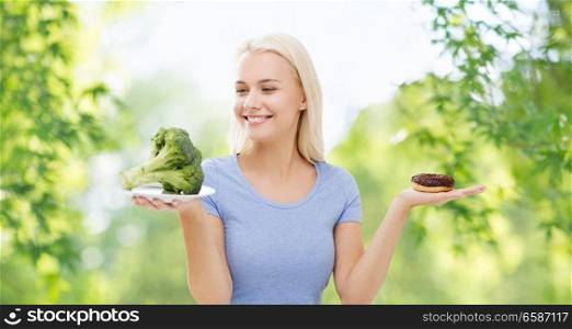 healthy eating, junk food, diet and choice people concept - smiling woman choosing between broccoli and donut over green natural background. smiling woman with broccoli and donut