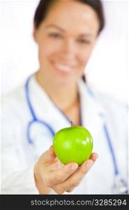 Healthy eating or lifestyle concept shot of a smiling woman doctor holding and offering a green apple, the focus is on the apple in the foreground.