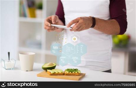 healthy eating, people and technology concept - close up of man with smartphone having vegetable sandwiches for breakfast at home kitchen over nutritional value chart. hands with phone and food nutritional value chart