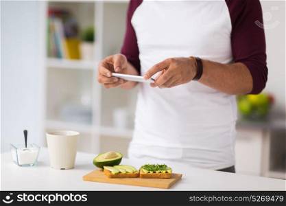 healthy eating, people and technology concept - man with smartphone having breakfast and photographing vegetable sandwiches at home kitchen. man photographing food by smartphone at home