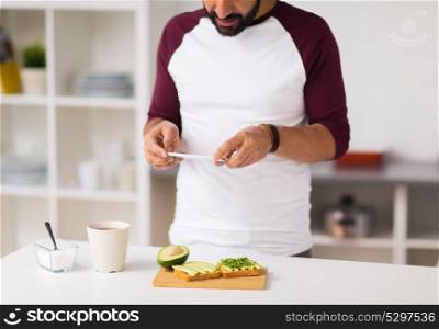 healthy eating, people and technology concept - man with smartphone having breakfast and photographing vegetable sandwiches at home kitchen. man photographing food by smartphone at home