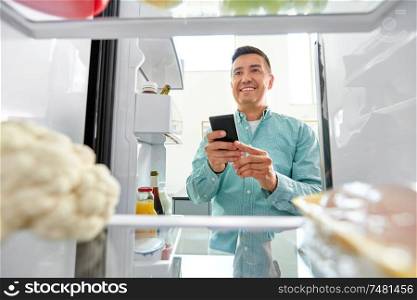 healthy eating, technology and diet concept - happy middle-aged man with smartphone making list of necessary food from fridge at home kitchen. man with smartphone making list of food in fridge