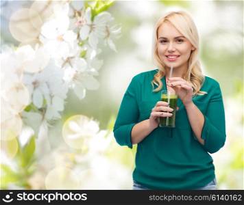 healthy eating, vegetarian food, diet, detox and people concept - smiling young woman drinking green vegetable juice or smoothie from glass over natural cherry blossom background