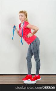 Healthy fit life style, controling body concept. Woman wearing sportswear, leggings and trainers standing on weight machine holding measuring tape.. Woman wearing sportswear standing on weight machine