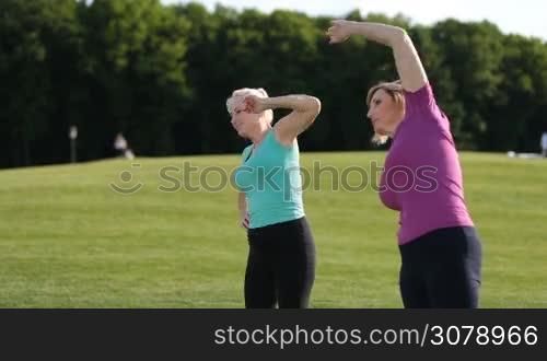 Healthy fitness senior blonde women doing side stretches and slopes in park. Side view. Positive smiling adult female friends warming up and doing stretching over colorful landscape background during workout outdoors on park lawn.