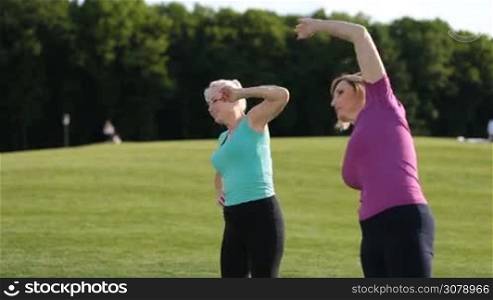 Healthy fitness senior blonde women doing side stretches and slopes in park. Side view. Positive smiling adult female friends warming up and doing stretching over colorful landscape background during workout outdoors on park lawn.