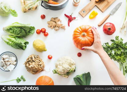 Healthy food and clean seasonal eating concept. Various organic vegetables on white desk background with pot , cutting board and knife. Female hand holding pumpkin. Vegetarian background. Top view