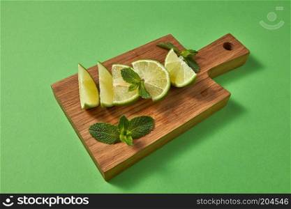 Healthy food diet concept. Slices of juicy ripe organic green lime, green mint on a wooden cutting board on a green background. Copy space.. Slices of fresh green lime and sprig of green mint on a wooden brown board on a green. Ingredient for homemade lemonade.
