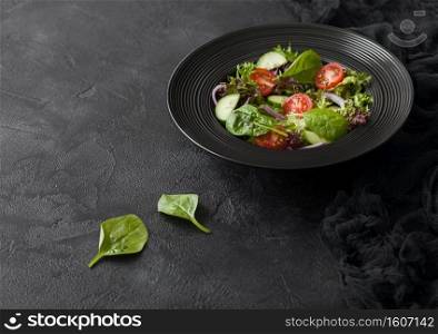 Healthy fresh vegetables salad with lettuce and tomatoes, red onion and spinach in black bowl on dark table background.