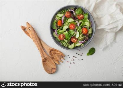 Healthy fresh vegetarian vegetables salad with lettuce and tomatoes, red onion and spinach in grey bowl plate on white table background with spatula fork and and spoon. Top view
