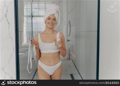 Healthy habits. Happy attractive slim woman enjoying morning routine at home, right after shower replenishes water balance with pure mineral glass of water and ready to measure herself with tape line. Happy attractive slim woman enjoying morning routine at home
