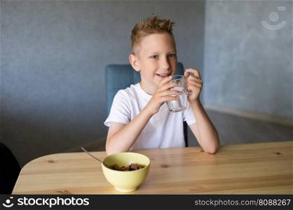 Healthy lifestyle, breakfast before school, cute boy, white T-shirt, collaboration, layout, advertising space, drinking water, cornflakes, dry breakfast, glass of water, sitting at the table. A cute boy drinks water and eats a dry breakfast at home and smiles