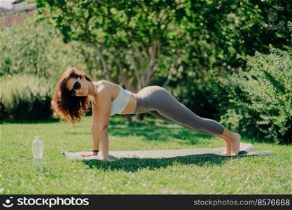 Healthy lifestyle concept. Horizontal shot of motivated fitness woman in active wear poses in plank pose on fitness mat exercises outdoors enjoys regular training wears sunglasses at summer time